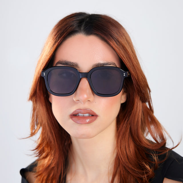 Woman with red hair in a black blouse wearing Bogota E Sunglasses Black with Grey Flat
