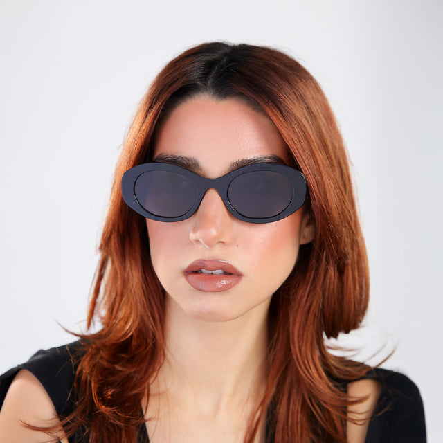 Women with red hair in a black blouse wearing Luna Sunglasses Black with Grey Flat