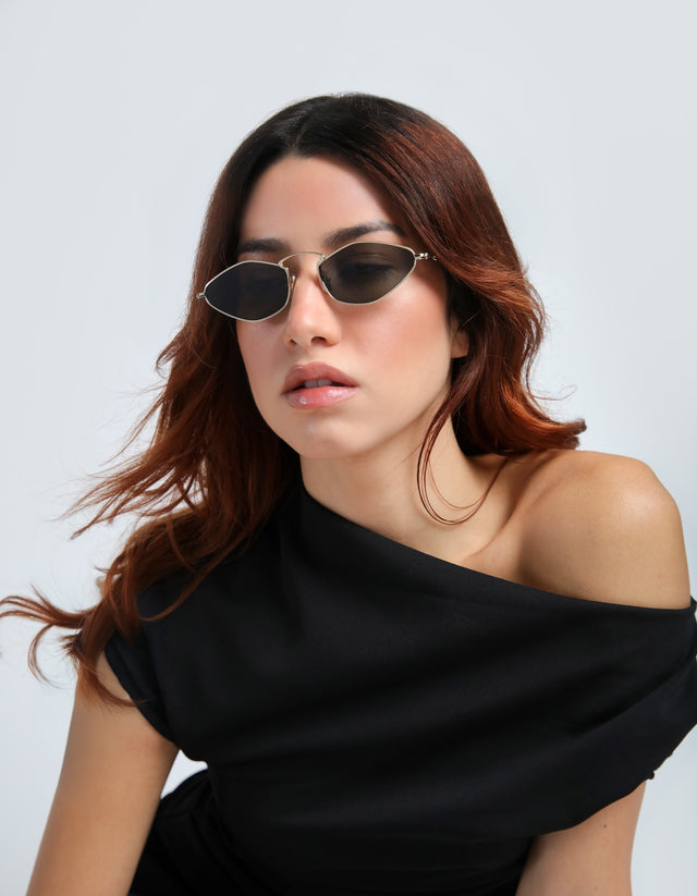 Woman wearing Austria sunglasses in Gold and a black off-shoulder top against a light gray background