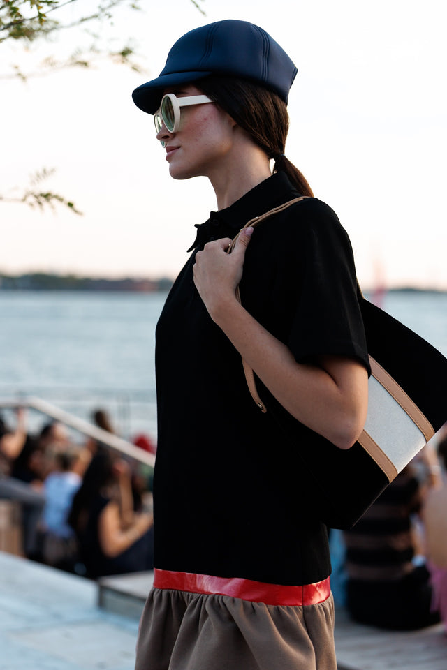 Woman in black top and cap holding a bag by New York waterfront wearing Cynthia Rowley  illesteva