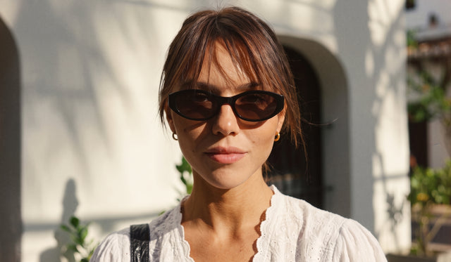 A woman wearing Bedford sunglasses in Black and a white textured top, outdoors against a sunlit white stucco wall