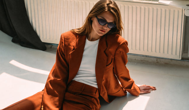 A woman wearing the Bedford Sunglasses and a rust-orange tailored blazer over a white tee, seated on the floor in front of a white radiator.