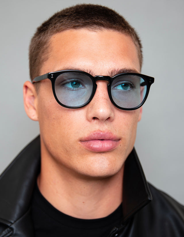 Model with buzzcut in a leather jacket wearing Eldridge in Black with custom blue lenses