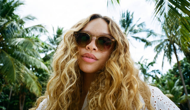 A woman with long, wavy blonde hair wearing round Leonard II Sunglasses in brown near palm trees