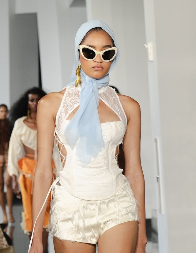 Model wearing Kim Shui x illesteva and a white outfit with a light blue scarf on a runway.