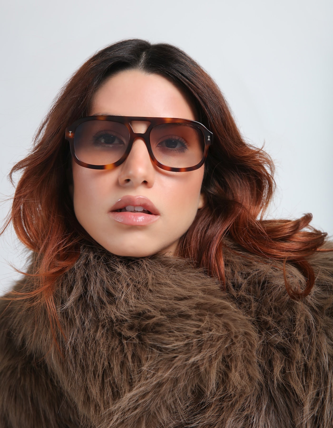 Woman wearing Memphis Sunglasses and a brown fur coat against a light gray background