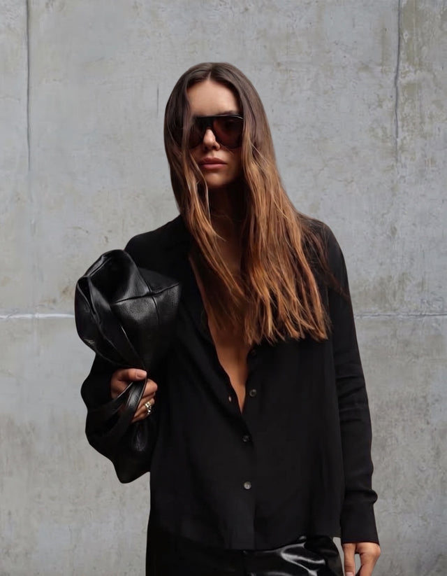 A woman with long brown hair wearing Telluride sunglasses, a black button-up shirt, and holding a black leather clutch bag near a concrete wall