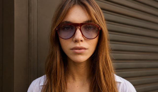 A close-up of a woman with long light brown hair wearing Vanderbilt sunglasses, set against a dark brown garage door.