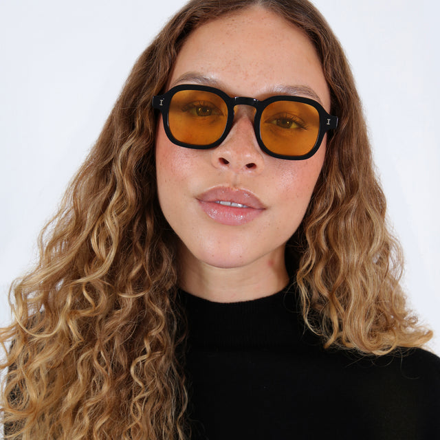 Brunette model with wavy hair in a black mockneck wearing Washington Sunglasses Black with Honey Flat See Through