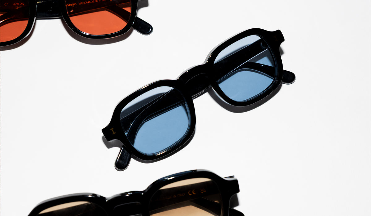 Three pairs of black sunglasses arranged on a clean white background: the center pair has blue-tinted lenses, while the top and bottom pairs have Sunset and Taupe lenses, respectively.