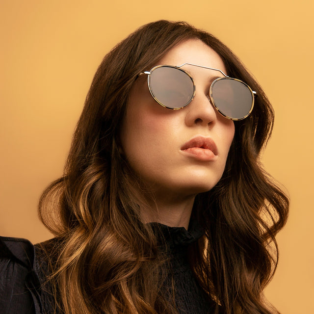 Brunette model in a ruffled, black dress wearing Wynwood Ace Sunglasses Tortoise/Silver with Grey Flat