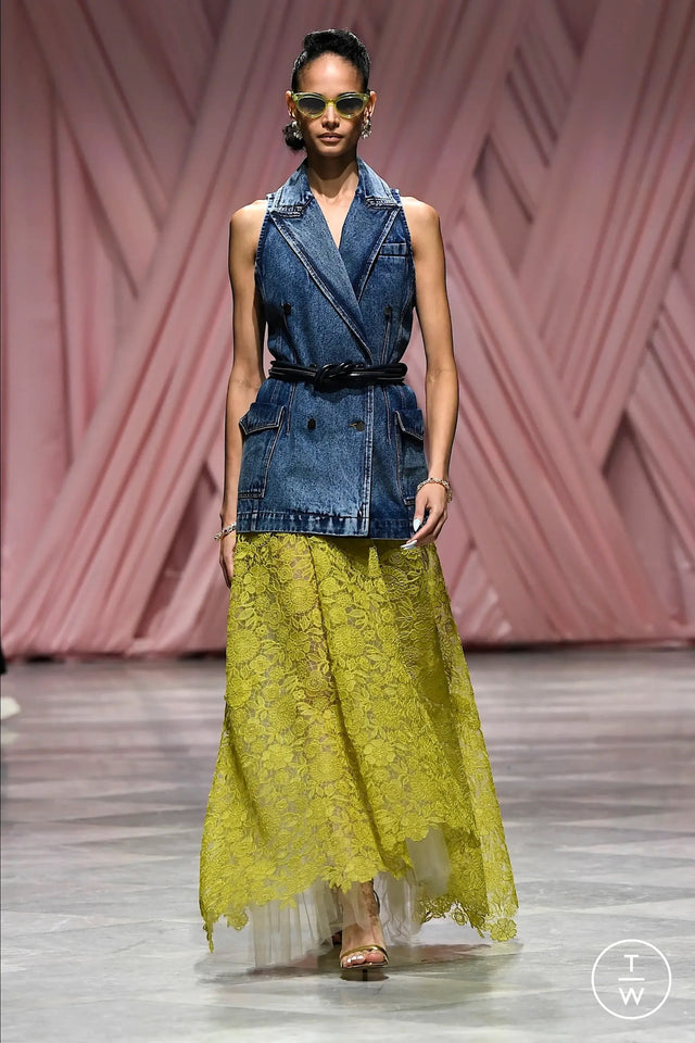 Model wearing a denim vest over a yellow lace dress and the Pamela Sunglasses on a runway.