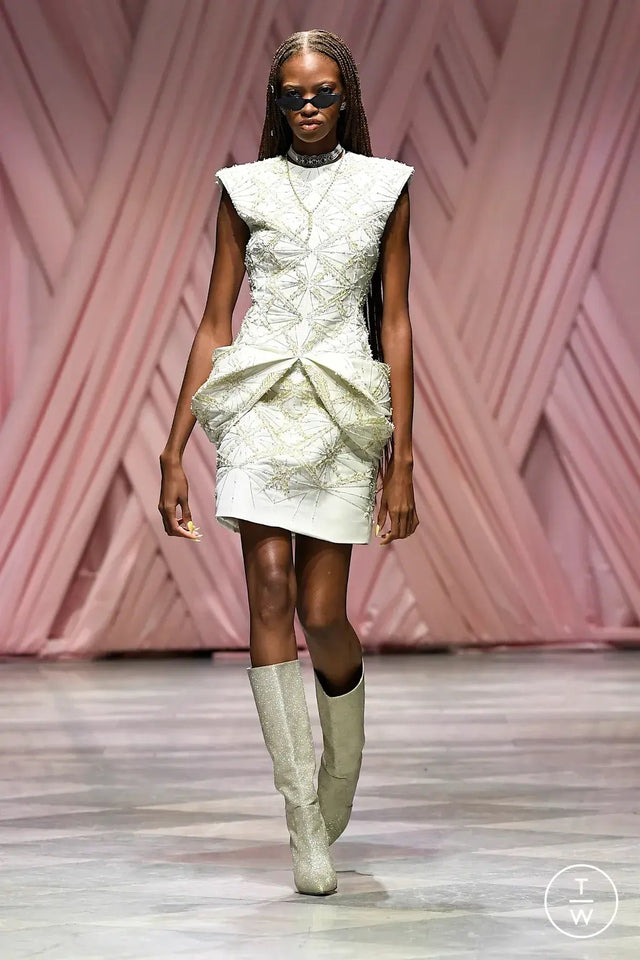 Model walking on a runway wearing a light-colored dress, boots, and the Marianne Sunglasses