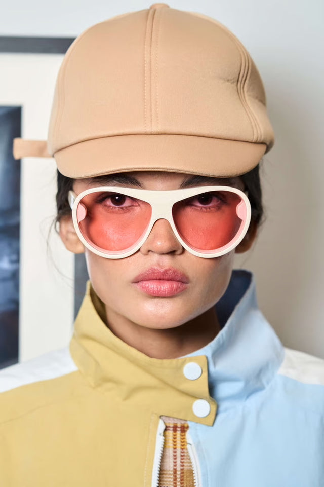 Close up of model in a cap wearing Cynthia Rowley x illesteva Sunglasses in Cream with Pink lenses